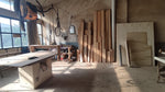 May include: A woodworking workshop interior featuring a large central workbench with a light-colored base, stacks of vertical wooden planks leaning against a wall, various tools hanging from the ceiling, and large windows letting in natural light.