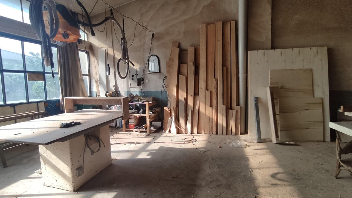 May include: A woodworking workshop with a large table, stacks of lumber, and various tools. The room has large windows and natural light, suggesting a space for crafting and construction.
