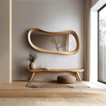May include: A modern interior scene featuring a large, organically shaped wooden mirror hanging above a wooden bench. A small vase with dried flowers and a woven ottoman are also present. The room has a neutral color palette with natural light.