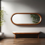 May include: A wooden-framed, oval mirror hangs above a bench with a cream-colored cushion. The mirror reflects greenery, and the bench has wooden legs. The room has hardwood flooring and a potted plant.