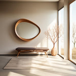 May include: A modern interior scene featuring a large, oval wooden mirror mounted on a neutral-toned wall. Below the mirror is a bench with a dark brown cushion and wooden legs. A vase with branches sits near a large window.