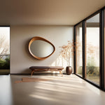 May include: A modern interior featuring a large, organic-shaped wooden mirror mounted on a beige wall. Below the mirror is a bench with a dark brown cushion. A potted plant and large windows complete the minimalist design.