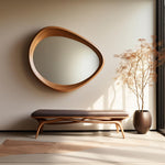 May include: A modern interior scene featuring a large, organic-shaped wooden mirror mounted on a neutral wall. Below, a wooden bench with a dark cushion complements the mirror. A potted plant adds a touch of nature to the space.