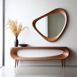 May include: A modern, wooden console table with a matching, asymmetrical mirror. The table has a curved design with four legs. A dark vase with dried flowers and a black bowl sit on the table. The mirror is a unique, organic shape.