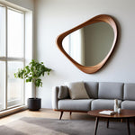 May include: A large, asymmetrical wooden mirror with a natural wood grain finish hangs on a white wall. The mirror's unique shape adds a modern touch to the room. A gray sofa, round coffee table, and potted plant complete the interior design.