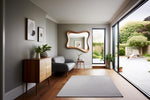 May include: A modern living room with a gray color scheme. Features include a wooden cabinet, a gray sofa, a large wavy-edged mirror, and a gray rug. A large glass door opens to a patio with greenery.