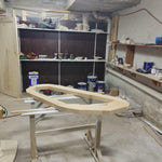May include: A light-colored, unfinished wooden boat frame rests on a white and beige sawhorse. The frame is in the shape of a boat hull. The background shows a workshop with shelves, paint cans, and other woodworking supplies.