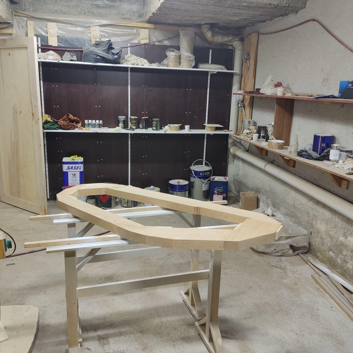 May include: A light-colored, unfinished wooden boat frame rests on a white and beige sawhorse. The frame is in the shape of a boat hull. The background shows a workshop with shelves, paint cans, and other woodworking supplies.