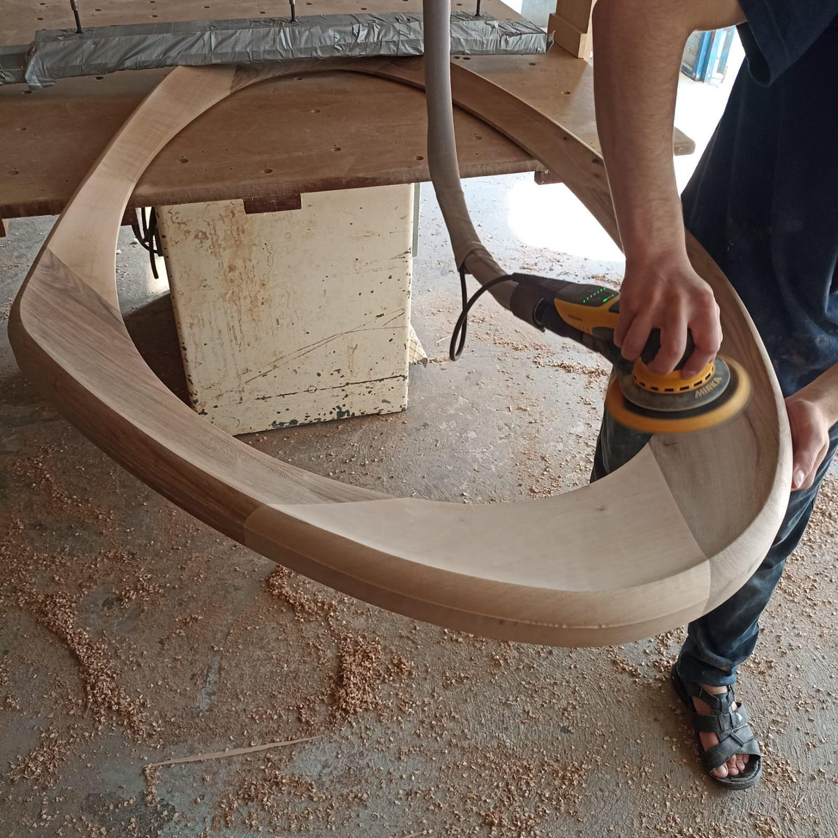 May include: A craftsman sands a large, unfinished wooden oval frame with a power sander. The frame is light brown and appears to be part of a larger furniture piece. Sawdust covers the floor, indicating a workshop setting.