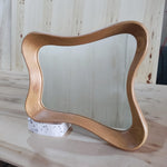 May include: A wooden mirror with a unique, wavy frame. The mirror is rectangular with rounded corners and is supported by a small, white, speckled block. The frame has a natural wood grain finish.