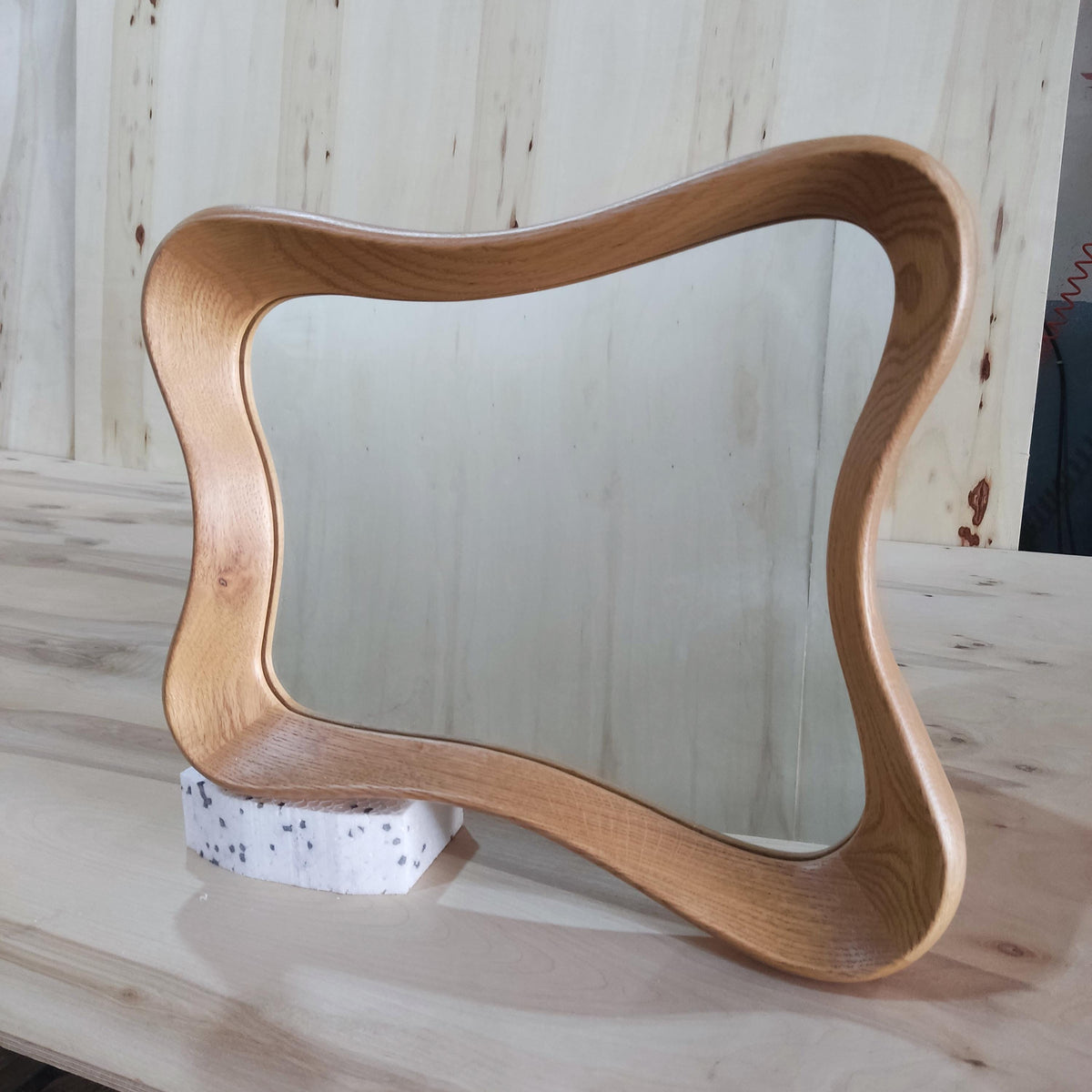 May include: A wooden mirror with a unique, wavy frame. The mirror is rectangular with rounded corners and is supported by a small, white, speckled block. The frame has a natural wood grain finish.