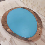 May include: Oval-shaped mirror with a teal-colored reflective surface. The mirror is framed with a dark wood border, featuring lighter wood accents at the corners. The wood grain is visible, adding a natural aesthetic.