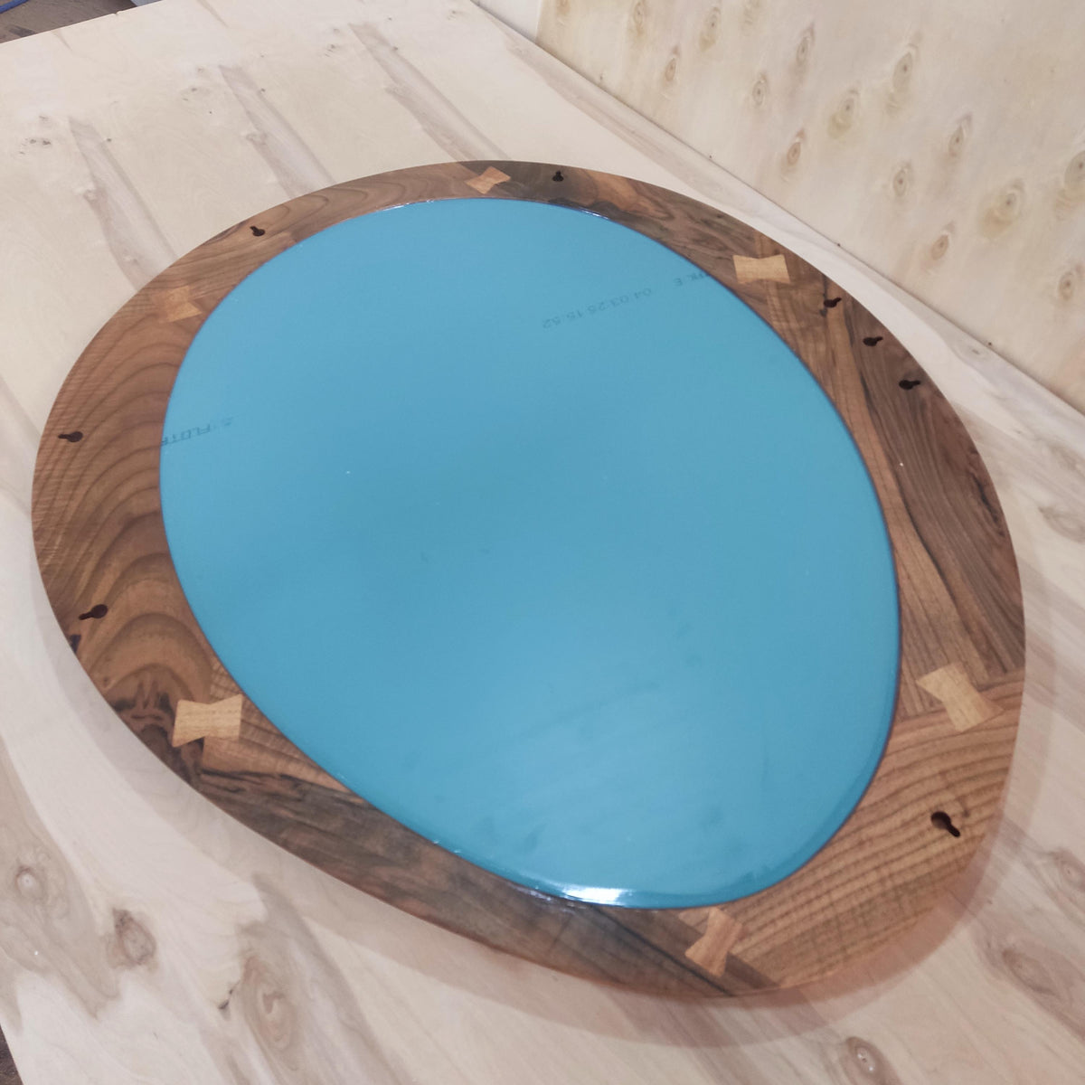 May include: Oval-shaped mirror with a teal-colored reflective surface. The mirror is framed with a dark wood border, featuring lighter wood accents at the corners. The wood grain is visible, adding a natural aesthetic.