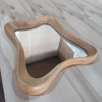 May include: A wooden mirror with a unique, wavy frame. The frame is made of light-colored wood and has a smooth, curved design. The mirror itself is rectangular and reflects the surrounding environment.