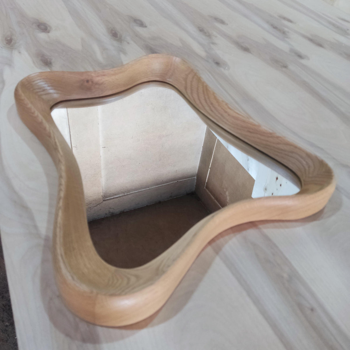 May include: A wooden mirror with a unique, wavy frame. The frame is made of light-colored wood and has a smooth, curved design. The mirror itself is rectangular and reflects the surrounding environment.