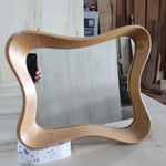 May include: A light-colored wooden mirror with a unique, wavy frame. The mirror is rectangular with rounded corners and sits on a white foam base. The wood grain is visible, and the mirror reflects the surrounding workshop.