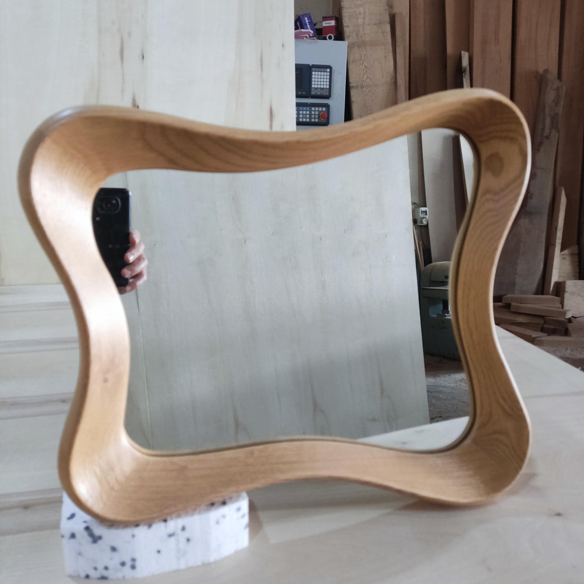 May include: A light-colored wooden mirror with a unique, wavy frame. The mirror is rectangular with rounded corners and sits on a white foam base. The wood grain is visible, and the mirror reflects the surrounding workshop.