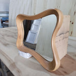 May include: A wooden mirror with a unique, wavy frame. The frame is made of light-colored wood and has a smooth, curved design. The mirror reflects a room with white doors and wooden surfaces. The mirror is resting on a white block.