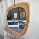 May include: A large, light-colored wooden mirror with a rounded rectangular shape. The frame is made of light-colored wood with a smooth, curved design. The mirror reflects a workshop setting.