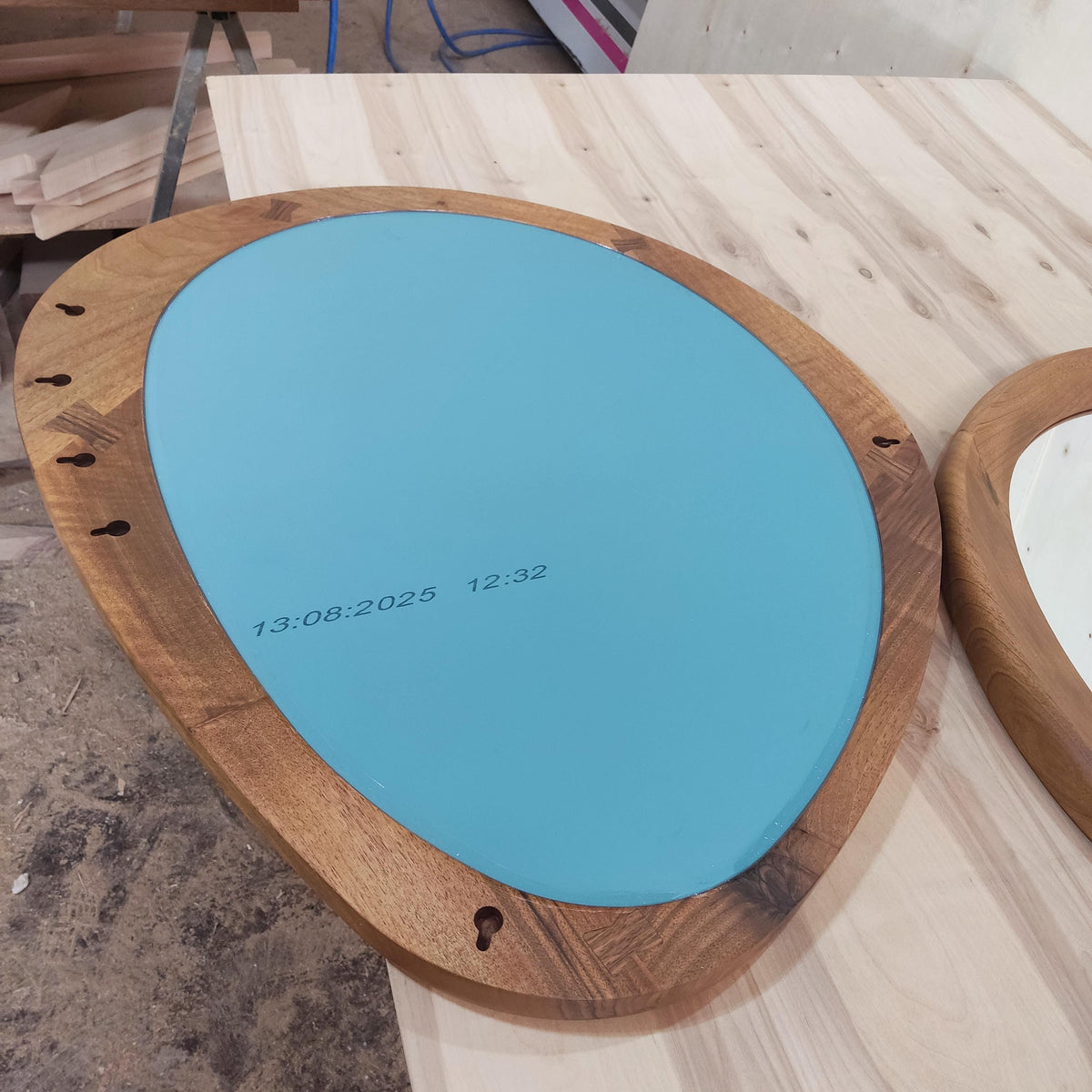 May include: Oval-shaped mirror with a light blue surface and a wooden frame. The frame is made of multiple wood pieces. The text "13:08:2025 12:32" is printed on the mirror's surface. The mirror has keyhole slots for hanging.