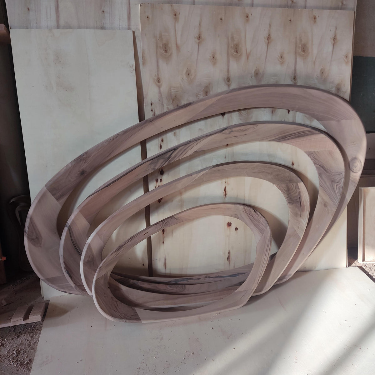May include: A set of four wooden oval-shaped frames, each nested within the other. The wood has a natural grain pattern with varying shades of brown and tan. The frames are arranged against a backdrop of wooden panels.