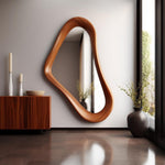 May include: A modern, organic-shaped wooden mirror hangs on a white wall. The mirror's frame has a warm, natural wood grain. A wooden cabinet, white candles, and a decorative bowl are visible in the room. The floor is a dark brown color.
