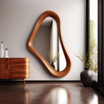 May include: A large, organically shaped wooden mirror hangs on a white wall. The mirror's frame is a warm, medium brown. The room features a wooden cabinet, vases, and a window with natural light.