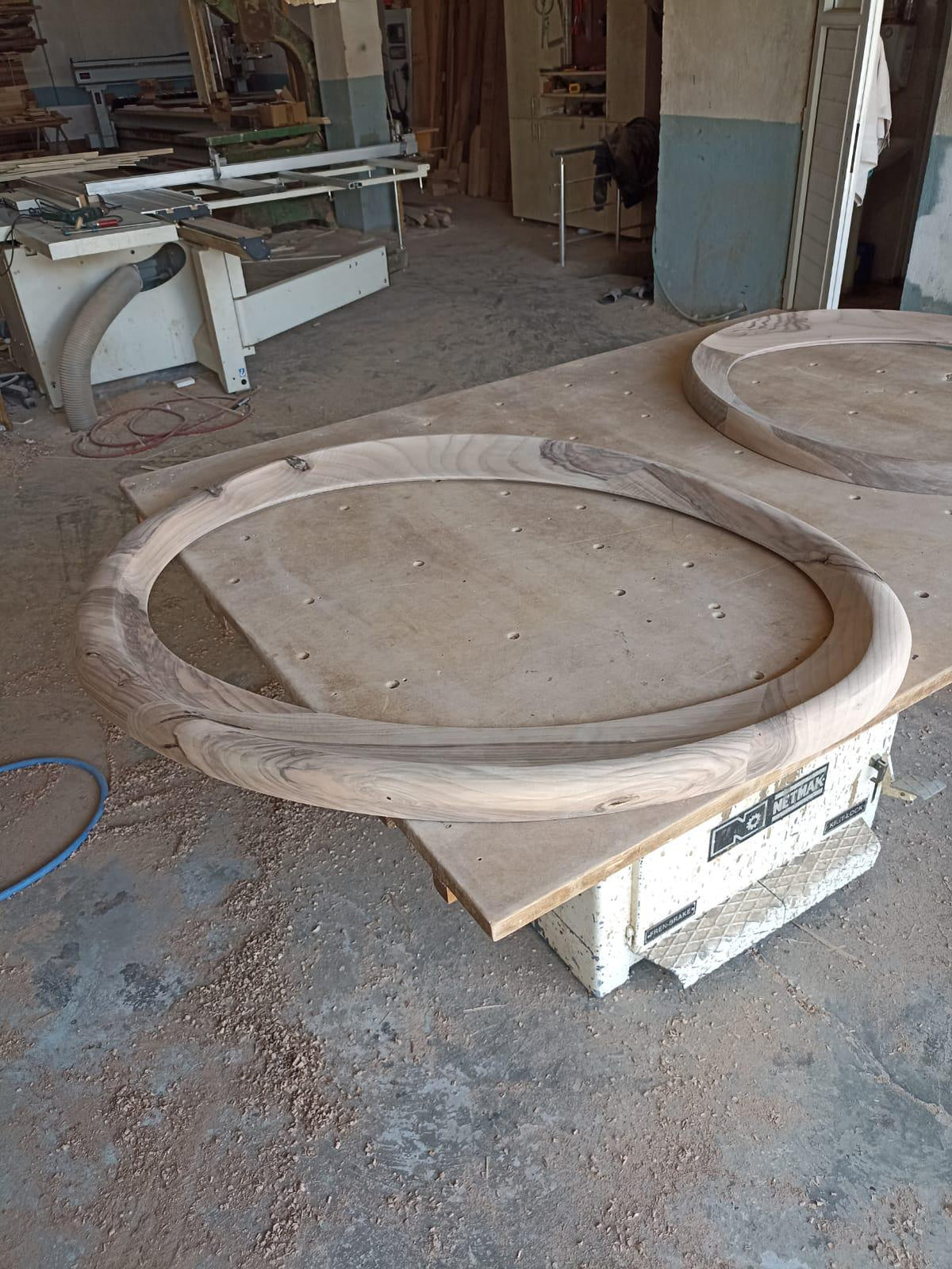 May include: Two unfinished wooden circular frames with a natural wood grain pattern sit on a workbench. The frames are thick and appear to be part of a larger woodworking project. The workshop setting includes tools and equipment.