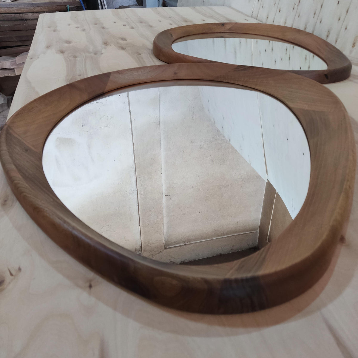May include: Two wooden mirrors with an organic, flowing shape. The frames are made of dark brown wood, and the mirrors have a clear reflective surface. The mirrors are placed on a light-colored wooden surface.