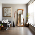 May include: A modern living room featuring a large, wooden, free-standing mirror with an organic shape. The room includes a gray sofa, a framed abstract artwork, and decorative vases with dried branches and greenery. The floor is made of wood.
