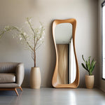 May include: A tall, wooden, full-length mirror with a unique, curved frame. The mirror is leaning against a neutral-colored wall. A decorative plant in a beige pot is next to the mirror. A sofa and another plant are also in the room.