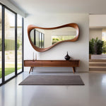 May include: A modern entryway featuring a large, organically shaped wooden mirror. Below, a long, low wooden console with three drawers and tapered legs holds decorative vases. A rectangular gray rug sits on the floor.