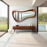 May include: A mid-century modern-style interior featuring a large, organically shaped wooden mirror. Below the mirror is a long, low wooden cabinet with a few decorative items. The room has large windows and a minimalist aesthetic.