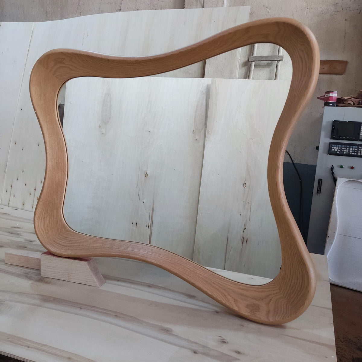 May include: A light brown, wooden, wavy-edged mirror frame. The frame has a unique, organic shape, supported by a small wooden base. The wood grain is visible, adding texture. The mirror frame is a decorative home decor item.