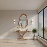 May include: A modern interior scene featuring a wooden console table with a curved base, topped with decor. Above, a uniquely shaped wooden mirror hangs on the wall. Two wall sconces flank the mirror. A potted plant and large window complete the room.