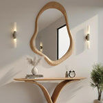 May include: A wooden, organically shaped mirror hangs above a wooden console table. The table holds a vase with dried flowers, books, a small clock, and a diffuser. Two wall sconces flank the mirror, and a small potted plant is on the right.