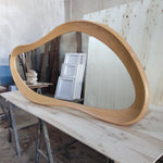 May include: A large, organically shaped mirror with a light brown wooden frame. The frame has a smooth, curved design. The mirror is resting on a light-colored wooden surface, reflecting the surrounding workshop environment.