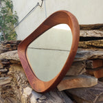 May include: A mid-century modern mirror with a unique, asymmetrical shape. The frame is crafted from warm-toned wood, and the mirror's surface reflects the surrounding environment. The design is minimalist and would complement a variety of interior styles.