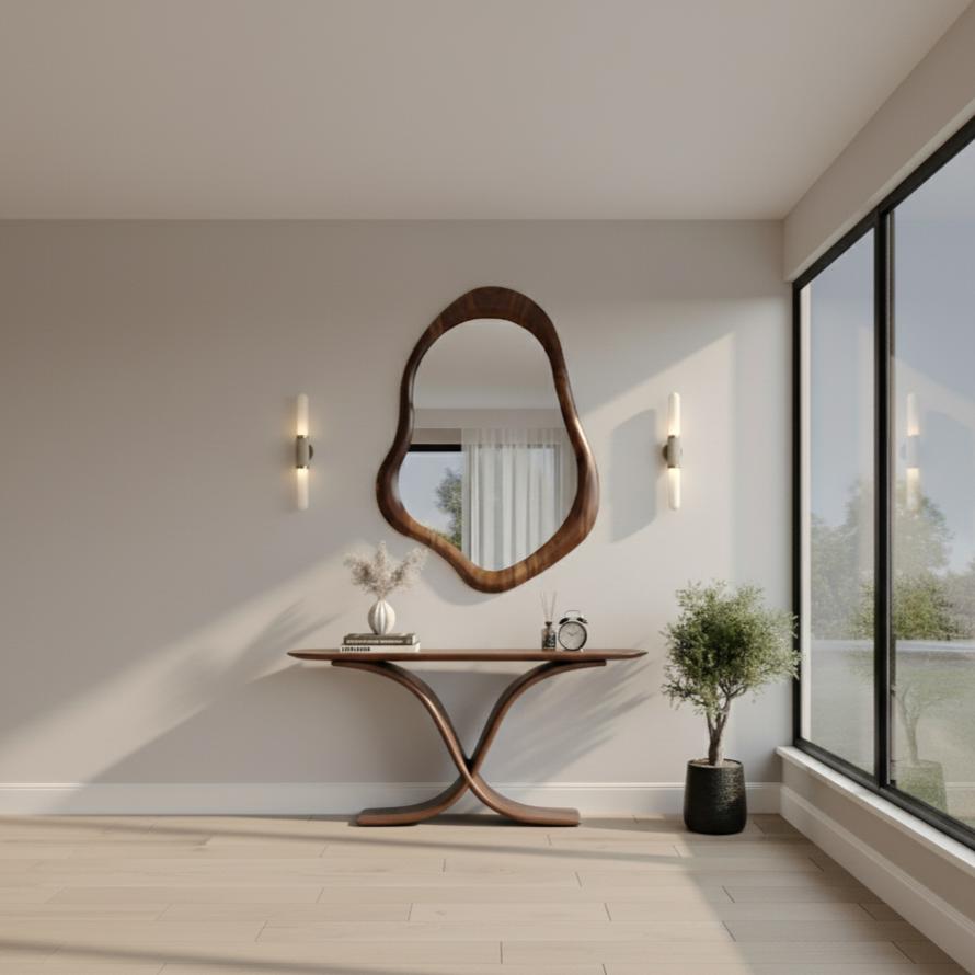 May include: A modern entryway featuring a large, organically shaped wooden mirror above a matching console table. The table has a unique crossed-leg design. Two sconces flank the mirror, and a small potted plant sits nearby. The room has light-colored walls and flooring.