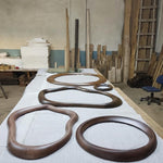 May include: Several wooden frames in various organic shapes and sizes are arranged on a white surface. The frames are a rich, dark brown color, with visible wood grain. A round mirror is also present. The background shows a workshop setting with wood planks.