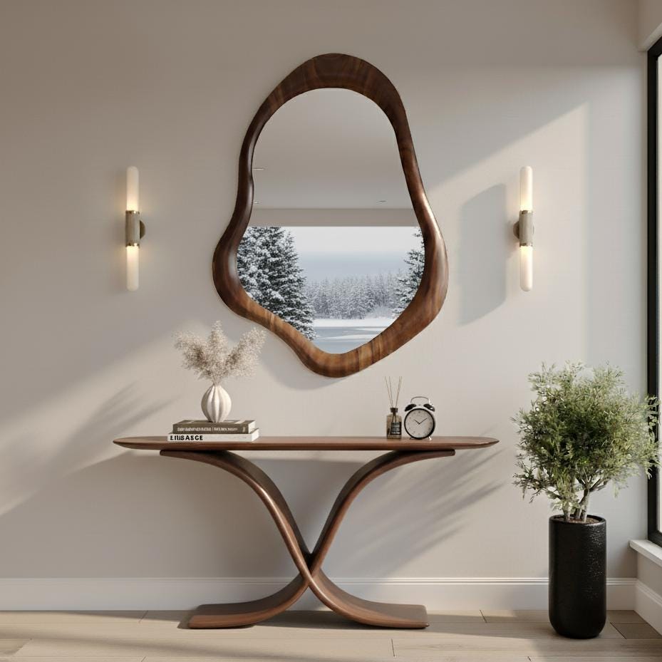 May include: A modern entryway scene featuring a large, organically shaped wooden mirror. Below, a matching console table with a curved base. The scene includes a small potted plant, a vase with dried flowers, and a decorative clock.