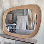 May include: A large, oval-shaped wooden mirror with a light brown, natural wood finish. The mirror has a thick, rounded frame and is displayed in a workshop setting. The mirror's shape is reminiscent of a rounded rectangle.