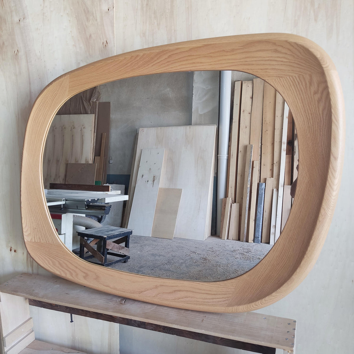May include: A large, oval-shaped wooden mirror with a light brown, natural wood finish. The mirror has a thick, rounded frame and is displayed in a workshop setting. The mirror's shape is reminiscent of a rounded rectangle.