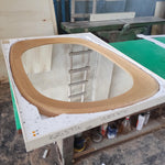 May include: Oval-shaped wooden framed mirror. The frame is made of light brown wood. The mirror is in a white protective container. The name "KRISTIN NELSON" is written on the container. The mirror is sitting on a green and brown table.