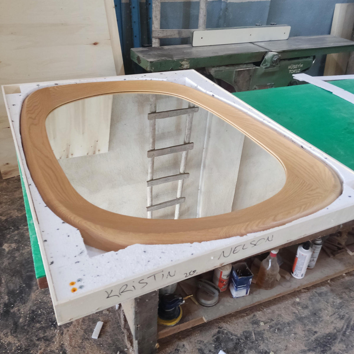 May include: Oval-shaped wooden framed mirror. The frame is made of light brown wood. The mirror is in a white protective container. The name "KRISTIN NELSON" is written on the container. The mirror is sitting on a green and brown table.