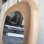 May include: A large, oval-shaped mirror with a light-colored, wooden frame. The frame has a smooth, curved design. The mirror reflects a workshop setting with tools and equipment. The wood grain is visible.