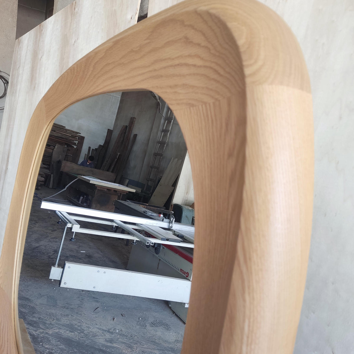 May include: A large, oval-shaped mirror with a light-colored, wooden frame. The frame has a smooth, curved design. The mirror reflects a workshop setting with tools and equipment. The wood grain is visible.