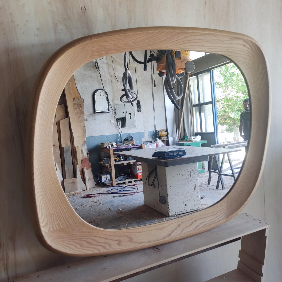 May include: A large, oval-shaped mirror with a light-colored wooden frame. The frame has a rounded, smooth design. The mirror reflects a workshop setting with tools and a person standing in the background. The wood grain is visible.