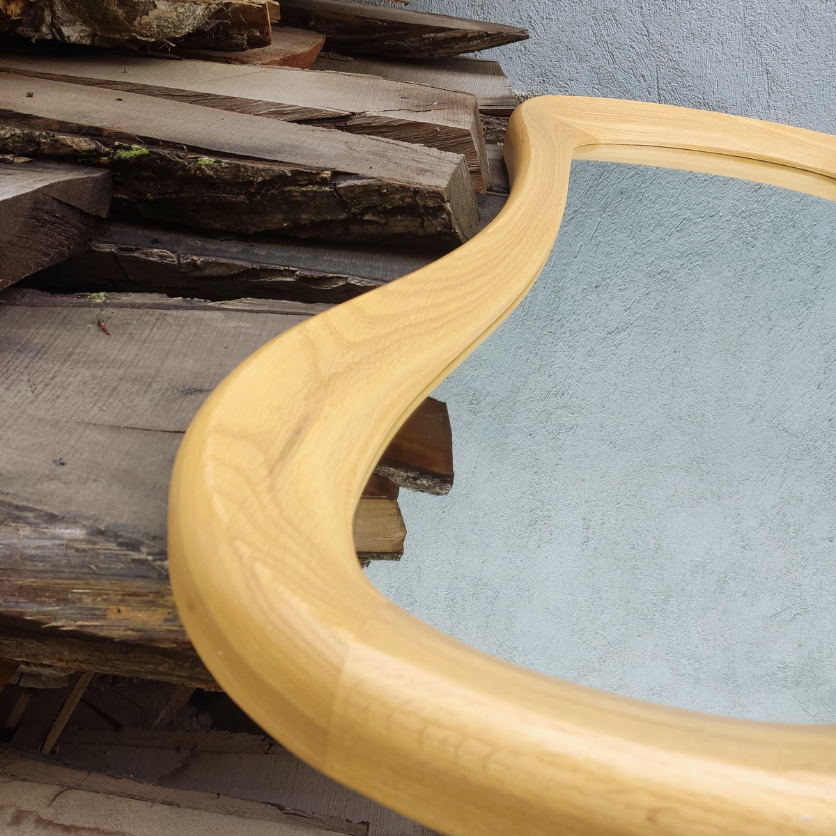 May include: A wooden framed mirror with a light-colored, curved frame. The mirror's reflective surface is visible, and the frame has a smooth, natural wood grain. The mirror is set against a background of stacked wooden planks.