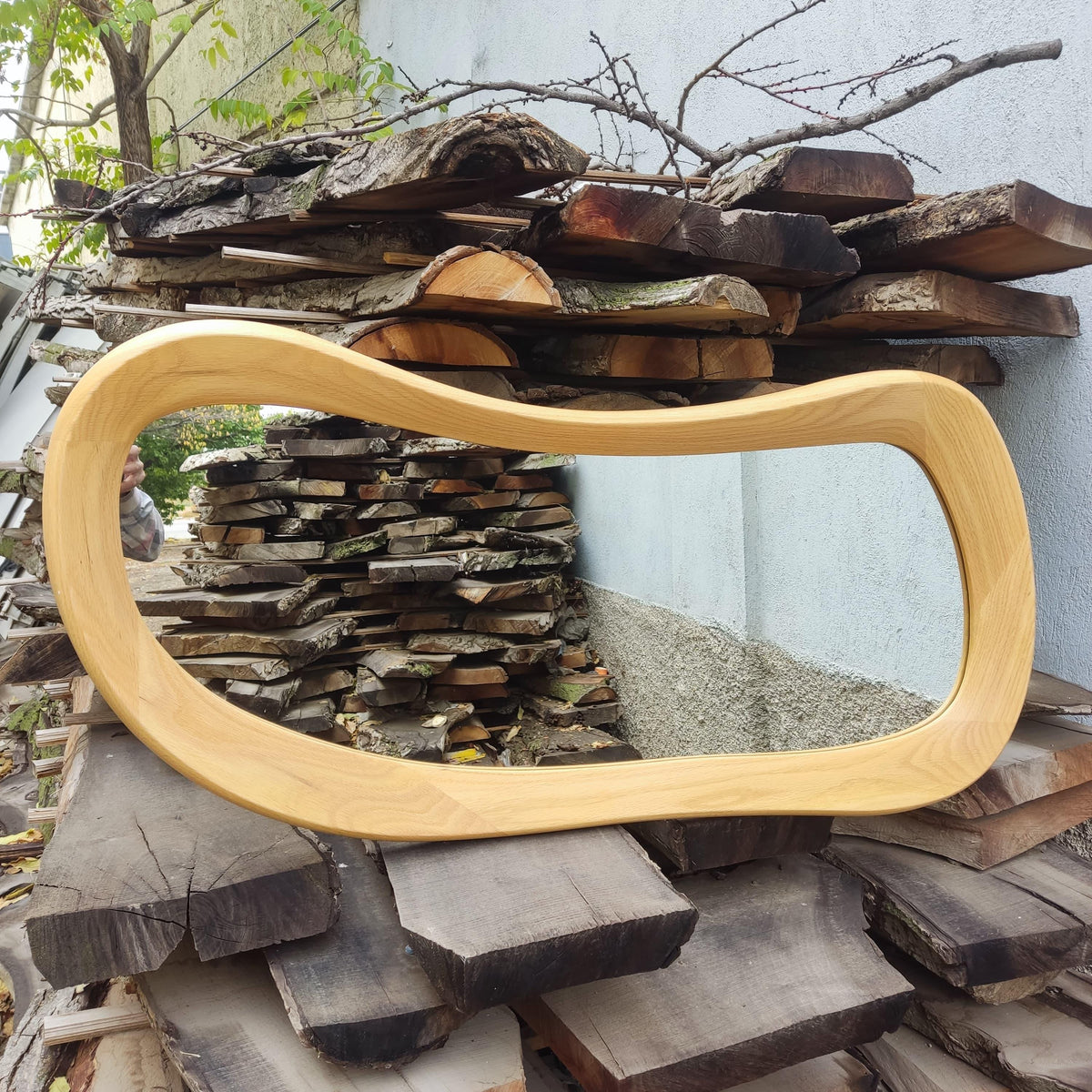 May include: A light-colored, wooden mirror with a unique, flowing, organic shape. The mirror's frame is made of smooth, light-toned wood. The mirror is displayed against a backdrop of stacked wooden planks and a light-colored wall.